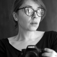 Anna Ciszek ilustratorka Zwierzobytów na zdjęciu trzyma aparat fotograficzny. Twarz skierowana prawym profilem z lekko uniesionymi brwiami znad okularów.
