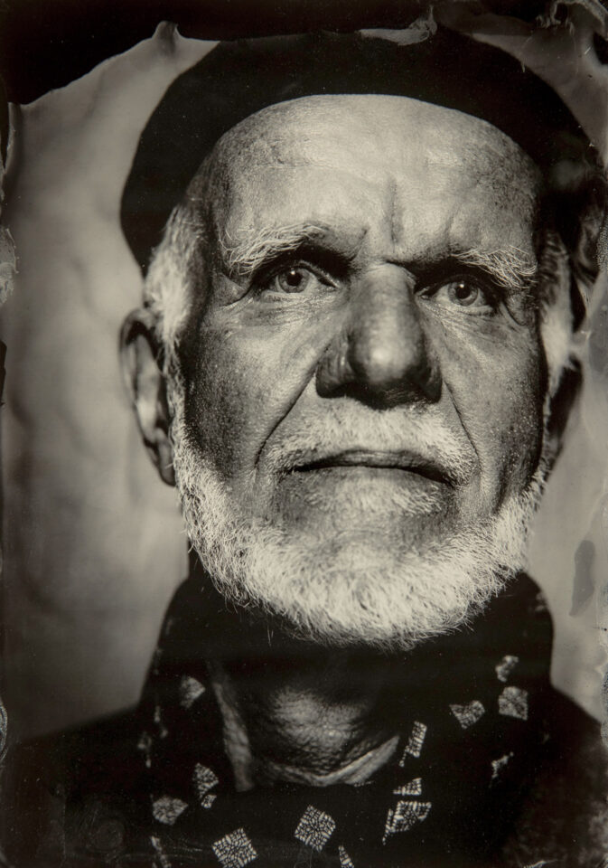 Portret Zbigniewa Bzdaka, 2023, Photo by Chris Walker. Starszy mężczyzna z siwym zarostem. Patrzy na wprost, ma szeroko otwarte oczy. Na głowie ma beret, przewiązany na szyi szal z printem w białe kwadraty. Zdjęcie jest w kolorze sepii, wykonane w technice mokrego kolodionu, starej techniki fotograficznyj. Charakteryzuje się ona dokładnością szczegółów. Na widać każdy włosek i zmarszczkę.