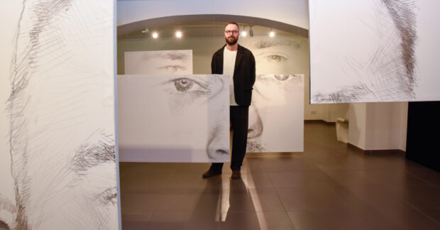 Artysta malarz Wiktor Jackowski stoi pośród swoich obrazów na wystawie Płaskoniż w Galerii Dolnej BWA w Kielcach.